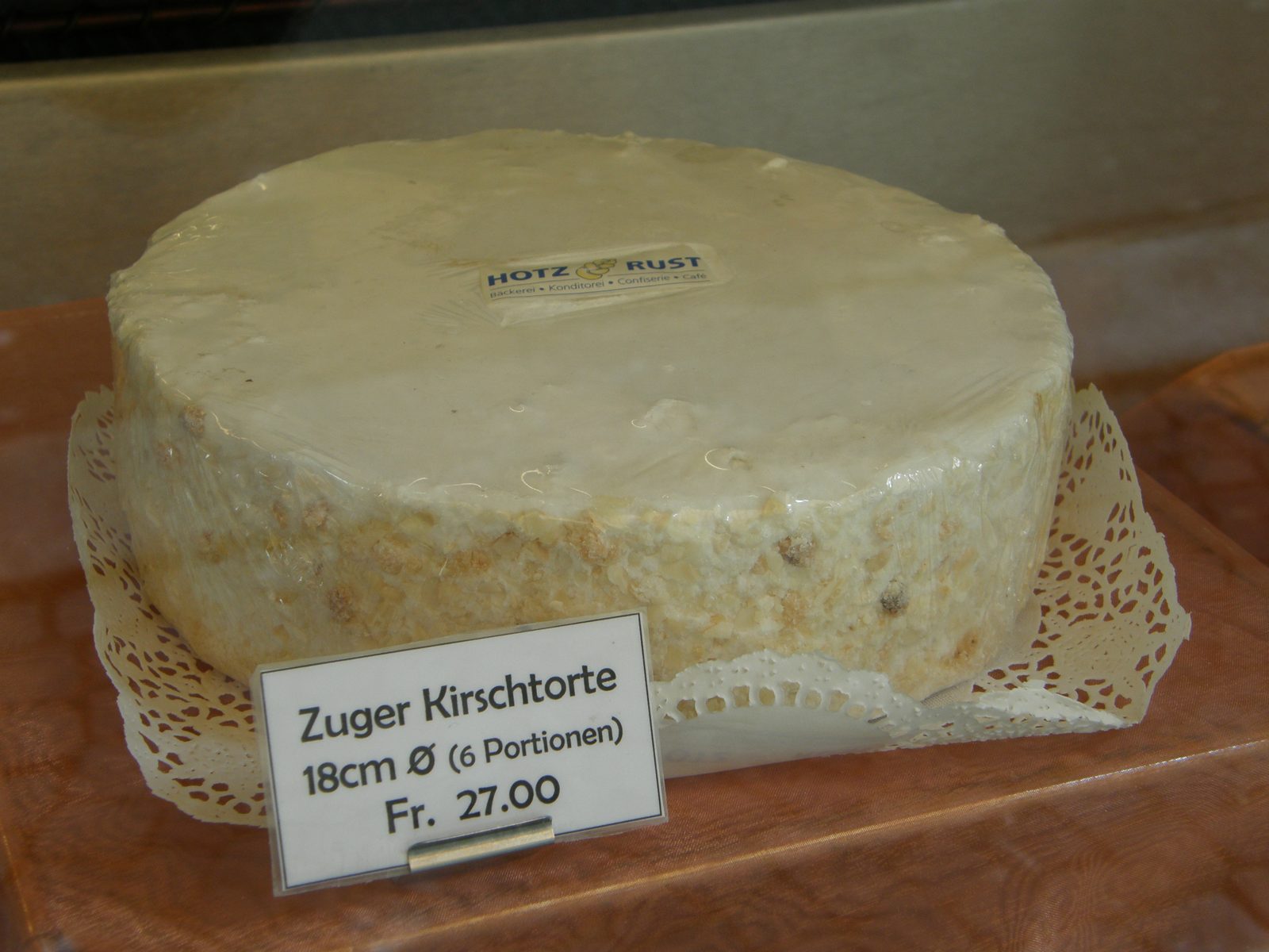 白い丸いケーキ。札に Zuger Kirschtorte 18cm Fr. 27.00