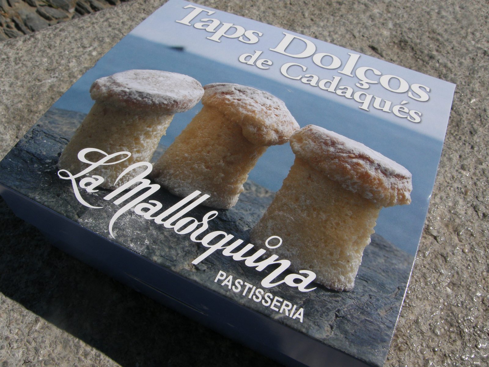 濃い青の箱に、海と岩の写真の上に立つきのこ形の焼き菓子三つ。La Mallorquina PASTISSERIA の文字