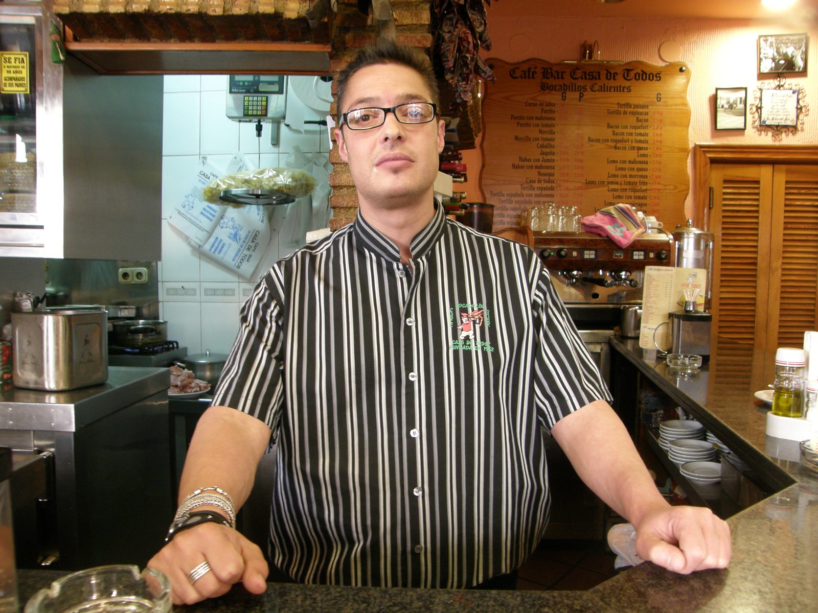眼鏡に縦じまのシャツの男性が、カウンターに両手をついて立つ。後ろに Café Bar Casa de Todos の品書き