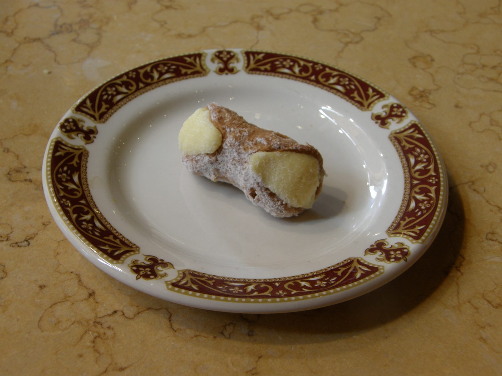 縁に模様のある白い皿に、粉砂糖をふった小さな筒形の菓子が一つ。両端から白いクリームがのぞき、手前の端に黄色いかたまりが付く
