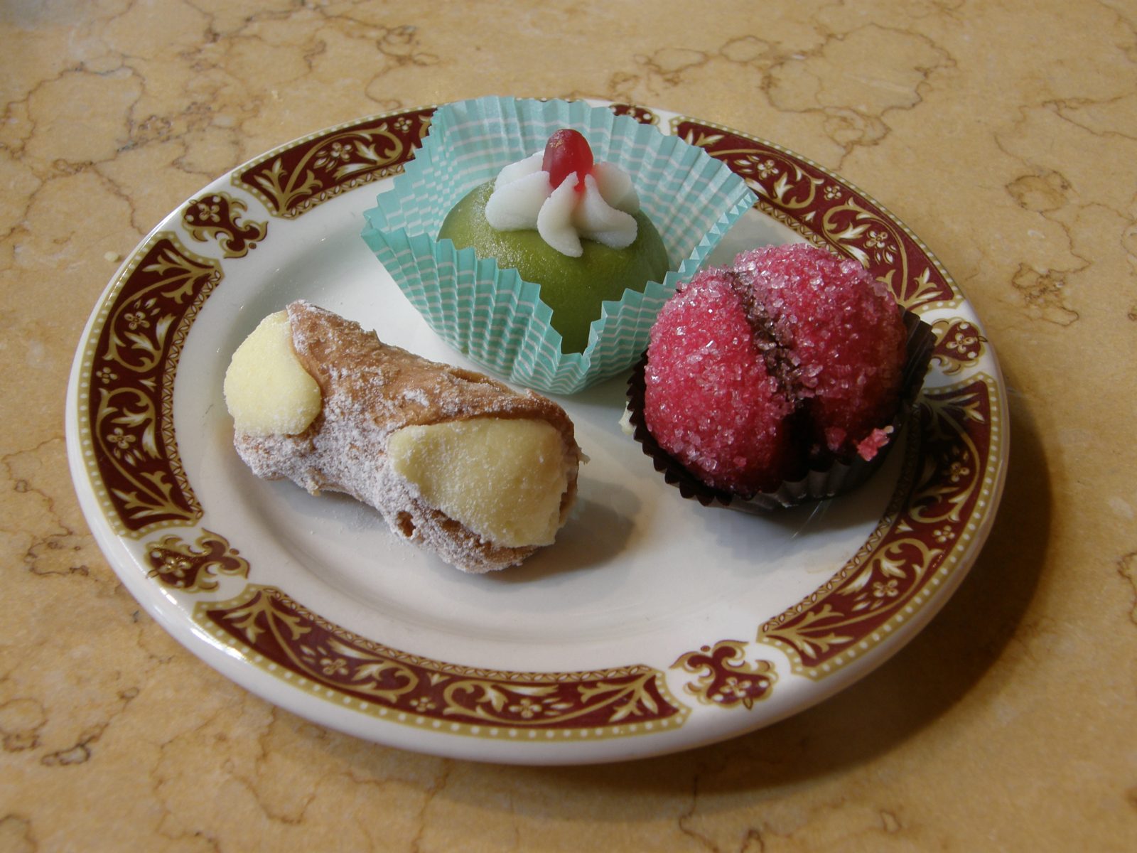 赤と金の縁の皿に、粉砂糖をふった筒形のカンノーロ、緑の小さなドーム形の菓子、赤い砂糖をまぶした丸い菓子がのる
