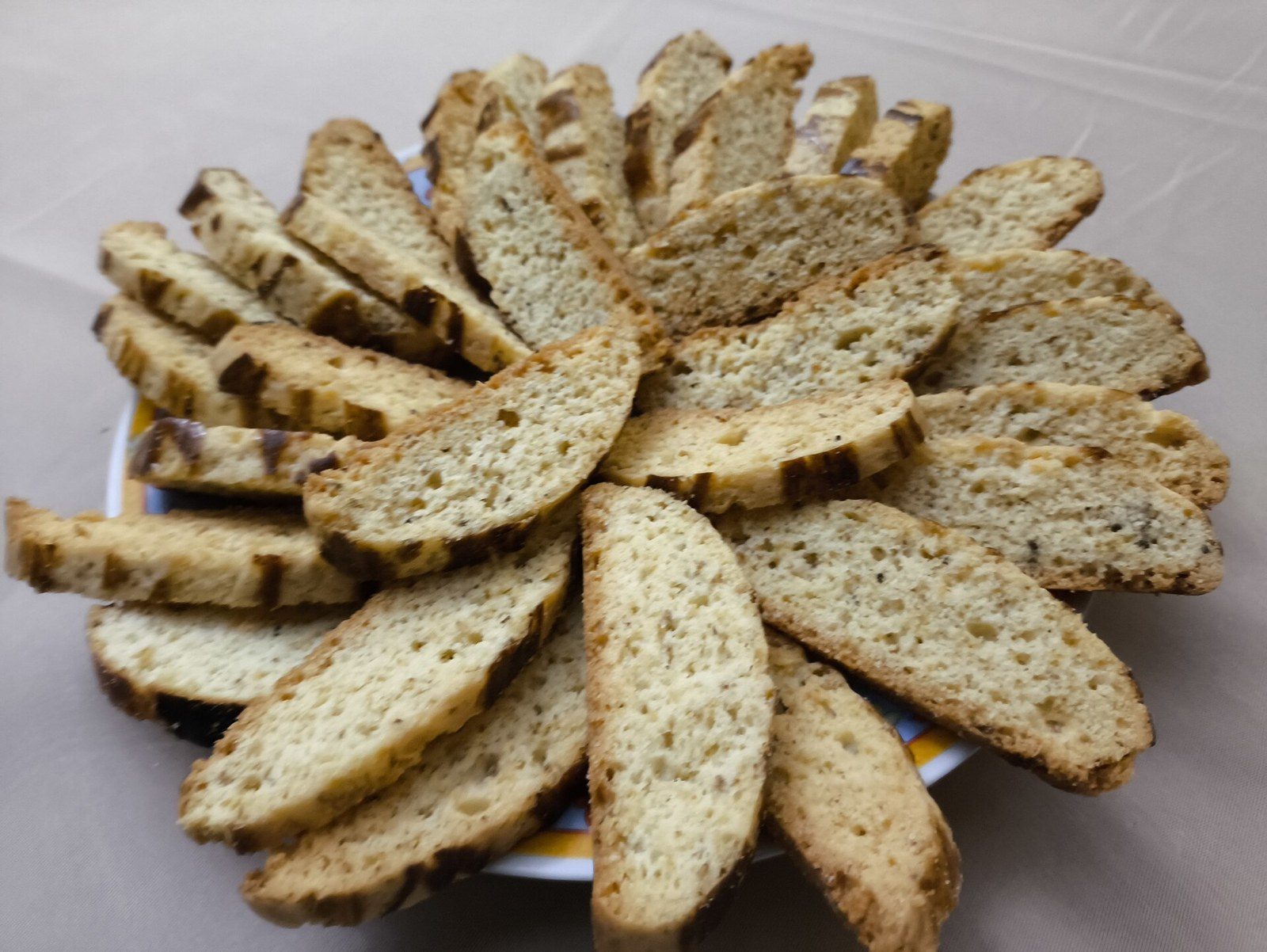 白い皿の上に、細長い薄切りの焼き菓子を放射状に並べている。切り口は淡い黄色で細かい粒が散り、縁に焼き色