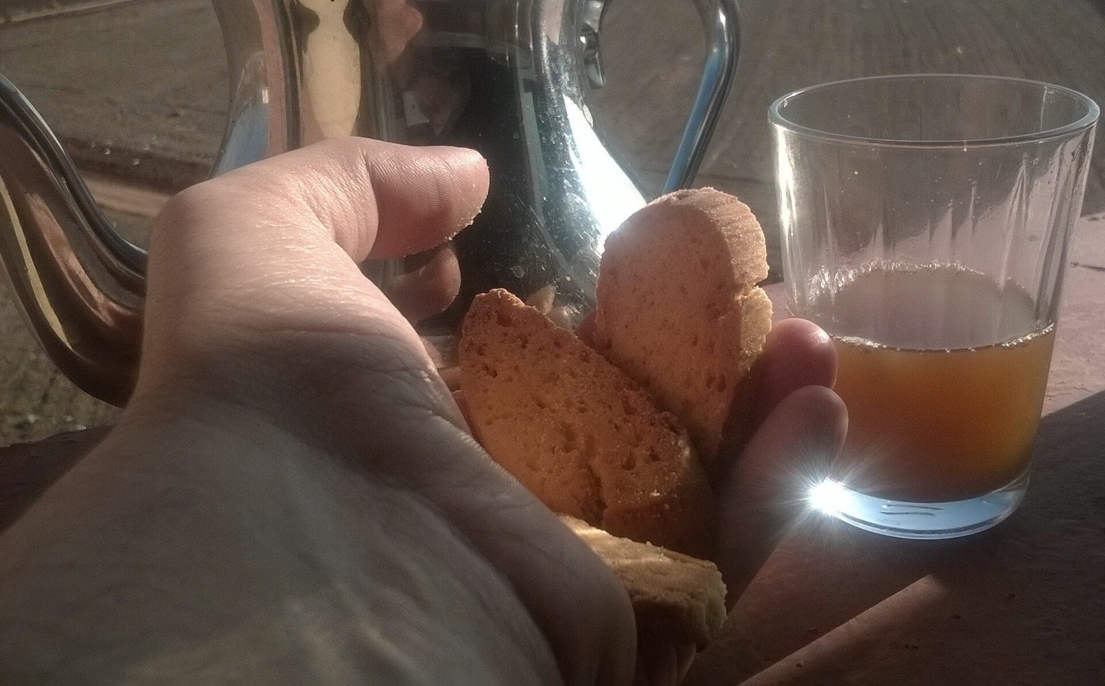 手のひらにのせた薄切りの焼き菓子。奥に銀色のティーポットと、茶の入ったガラスのコップ