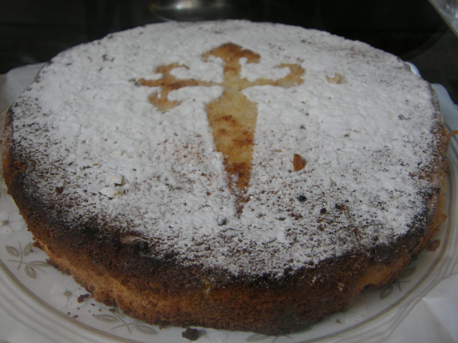 白い皿にのった丸いケーキ。上一面に粉砂糖がかかり、真ん中に下が細くとがった十字の形だけ茶色い焼き色が見える